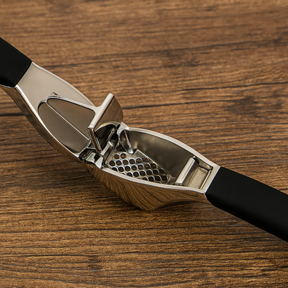 Presse A ail inox ouvert, montrant la trémie et la grille à trous ronds sur un plan de travail en bois.
